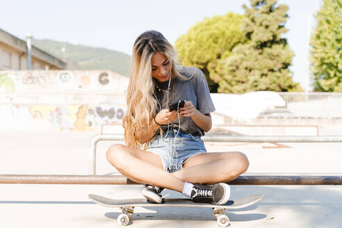 Blonde Frau, die ihr Smartphone benutzt, während sie mit dem Skateboard im Park an einem sonnigen Tag sitzt - FMOF01178