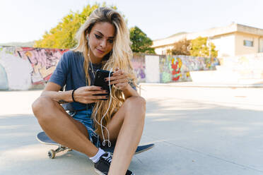 Junge blonde Frau, die ein Smartphone benutzt, während sie mit einem Skateboard im Park sitzt - FMOF01179