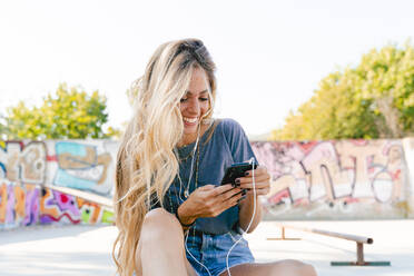 Glückliche junge blonde Frau, die ihr Smartphone benutzt, während sie mit einem Skateboard im Park sitzt - FMOF01180