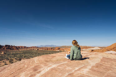 Frau sitzt auf einer Felsformation im Valley of Fire State Park vor blauem Himmel, Nevada, USA - DGOF01538