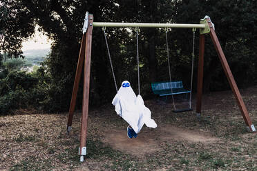 Unrecognizable kid in white ghost costume swinging and playing on playground during Halloween holiday - ADSF15876