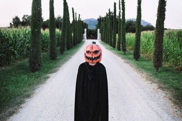 Anonyme Person mit gruseliger Halloween-Kürbismaske und schwarzem Mantel auf sandiger Straße stehend - ADSF15877