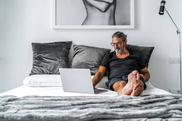 Smiling middle aged male entrepreneur sitting on bed and typing on laptop while working remotely on online project - ADSF15891