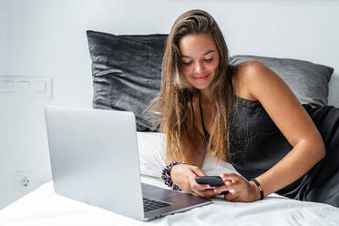 Positive Frau in Nachtwäsche, die auf einem weichen Bett liegt und am Morgen nach dem Schlaf Nachrichten auf dem Smartphone liest - ADSF15892