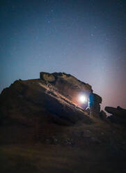 Anonymous tourist standing with illuminated torch on stone in mountains on background of starry sky at night - ADSF15905