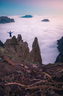 Rückenansicht eines anonymen männlichen Reisenden, der mit ausgestreckten Armen auf einem felsigen Hügel im Hochland steht und die herrliche Aussicht auf die Wolken genießt - ADSF15916