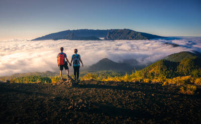 Rückenansicht des zärtlichen reisenden Paares, das auf einem Hügel steht und die Hände hält, während es die spektakuläre Landschaft auf den Wolken über dem bergigen Gelände bewundert - ADSF15928