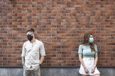 Trauriges Paar mit Gesichtsmasken, das während der Coronavirus-Krise an einer Mauer steht - EYAF01341