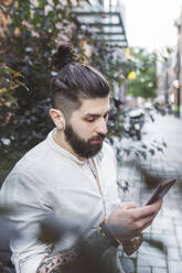 Hipster-Mann, der sein Smartphone benutzt, während er neben Pflanzen steht - EYAF01347