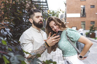 Boyfriend gesturing while showing smart phone to girlfriend on footpath in city - EYAF01349