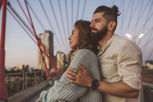 Nachdenkliches Paar, das bei Sonnenuntergang auf einer Brücke in der Stadt steht und wegschaut - EYAF01358