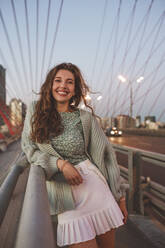 Lächelnde Frau steht auf einer Brücke gegen den Himmel in der Stadt bei Sonnenuntergang - EYAF01361