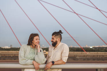 Lächelnder Freund und Freundin stehen auf einer Brücke gegen den Himmel in der Stadt bei Sonnenuntergang - EYAF01362