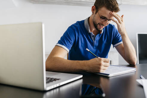 Smiling handsome man reading from book while sitting with laptop at home - JRFF04729