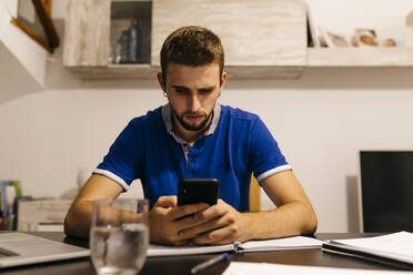 Gut aussehender junger Mann, der ein Smartphone benutzt, während er mit einem Arbeitsbuch am Tisch sitzt - JRFF04747