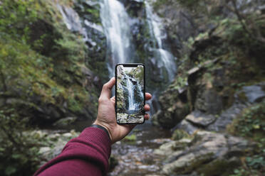 Die Hand eines Mannes hält ein Smartphone, während er die berühmte Fervenza do Toxa im Wald fotografiert, Provinz Pontevedra, Galicien, Spanien - RSGF00297