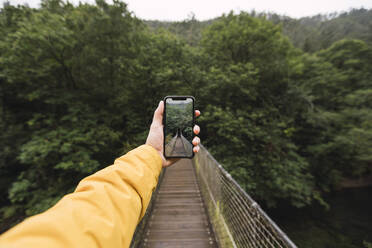 Hand eines Mannes, der sein Smartphone hält, während er eine Fußgängerbrücke vor Bäumen fotografiert, Fragas do Eume, Galicien, Spanien - RSGF00304