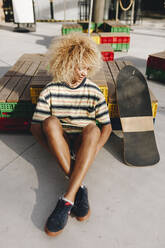 Afro blonde Frau sitzt am Skateboard an einem sonnigen Tag - MRRF00506