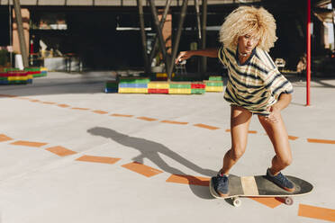 Selbstbewusste blonde Frau beim Skateboardfahren auf dem Fußweg an einem sonnigen Tag - MRRF00513