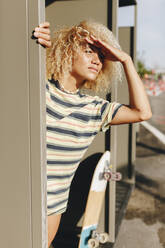 Blonde Afro-Frau, die ihre Augen abschirmt, während sie von einer Metallwand an einem sonnigen Tag wegschaut - MRRF00518