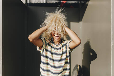 Playful woman with hands in curly blond hair amidst metallic walls on sunny day stock photo