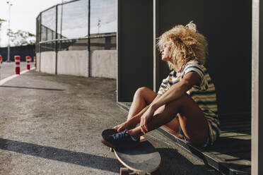 Nachdenkliche Frau mit Skateboard sitzend und wegschauend an einem sonnigen Tag - MRRF00525