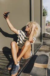 Blond woman taking selfie through smart phone while sitting against metallic wall - MRRF00526