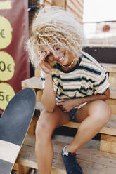 Lächelnde blonde Afro-Frau sitzt mit Skateboard auf Holzstufen - MRRF00534