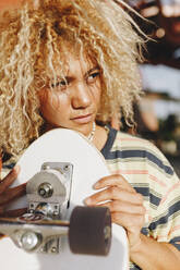 Hispanische Frau mit blonden lockigen Haaren, die wegschaut, während sie ein Skateboard an einem sonnigen Tag hält - MRRF00538