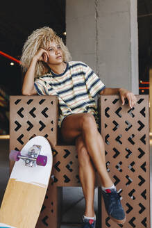Nachdenkliche blonde Afro-Frau sitzt mit gekreuzten Beinen - MRRF00544