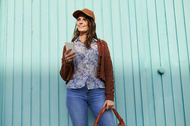 Young woman using mobile phone while standing against blue metal door - KIJF03289