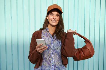 Lächelnde junge Frau, die ihre Handtasche hält, während sie ihr Mobiltelefon gegen eine blaue Metalltür hält - KIJF03290