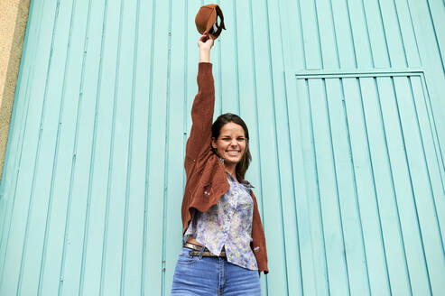 Happy woman with hand raised holding cap up while standing against blue metal door - KIJF03295