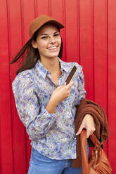 Lächelnde Frau, die ihre Jacke und ihre Handtasche hält, während sie ein Mobiltelefon gegen eine rote Metalltür hält - KIJF03301