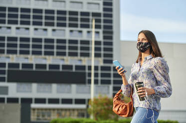Young woman wearing face mask using mobile phone while walking in city - KIJF03304