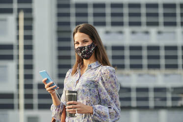 Young woman wearing face mask holding thermos while using mobile phone in city - KIJF03305