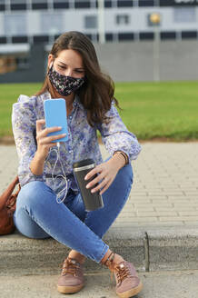 Junge Frau mit Gesichtsmaske, die ein Mobiltelefon benutzt, während sie auf einem Fußweg in der Stadt sitzt - KIJF03312