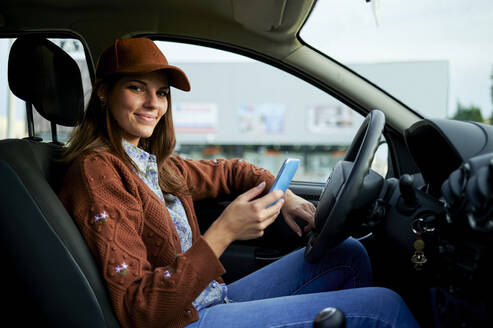 Lächelnde junge Frau, die ein Mobiltelefon benutzt, während sie im Auto sitzt - KIJF03324