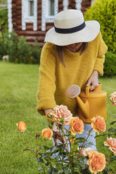 Frau mit Hut, die im Gewächshaus stehend eine Rose untersucht - KNTF05496