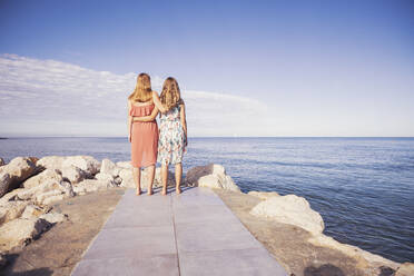 Back view of anonymous female friends in swimwear and summer clothes  embracing while standing on stony coast against waving sea during vacation  together in Puerto Escondido stock photo