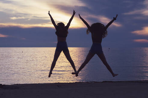 Freunde springen bei Sonnenuntergang mit erhobener Hand gegen das Meer - DHEF00447