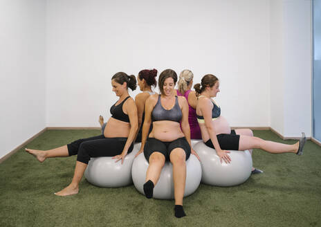 Schwangere Frau sitzt im Yoga-Studio auf einem Fitness-Ball in einem runden Kreis mit hochgelegten Beinen - MPPF01126