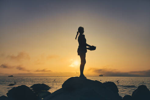 Silhouette einer Frau, die eine Schüssel hält, während sie bei Sonnenuntergang auf einem Felsen am Strand steht - KNTF05508