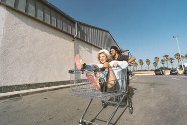 Young man pushing girlfriend sitting in shopping cart outside supermarket - DAMF00506