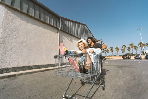 Junger Mann schiebt seine Freundin im Einkaufswagen vor einem Supermarkt - DAMF00506