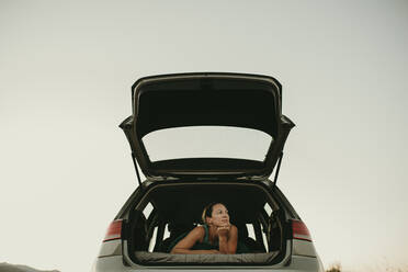 Thoughtful woman looking away while lying in car trunk against clear sky - DMGF00145
