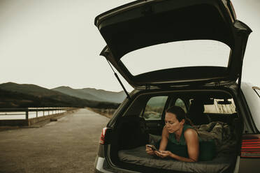 Frau, die ihr Smartphone benutzt, während sie im Kofferraum eines Autos liegt, gegen den klaren Himmel - DMGF00146