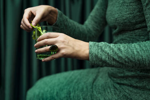 Midsection of woman holding glass of cocktail drink sitting against curtain stock photo