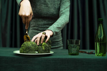 Mittelteil einer Frau, die Brokkoli mit einer Gabel in einem Teller auf dem Tisch schneidet - ERRF04525
