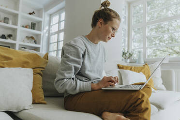 Mid adult woman working on laptop while sitting on sofa at home - MFF06198